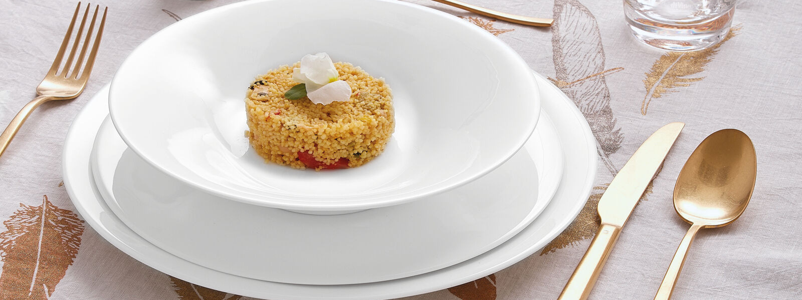 Piatto fondo bianco con cous cous e fiore decorativo, posate rame e oro su tovaglia elegante, apparecchiatura raffinata